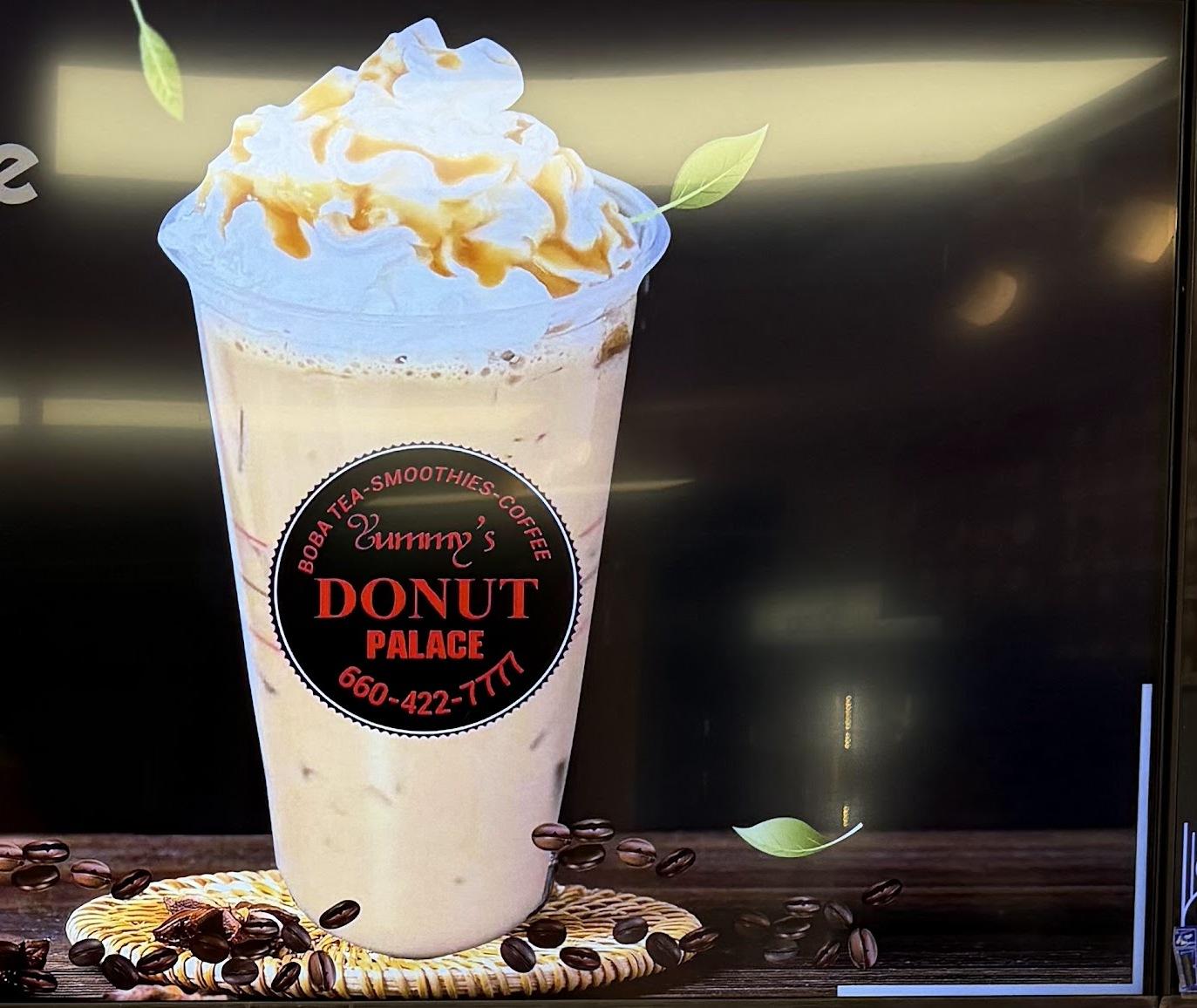 The backlit menu board at Yummy's: a tall iced coffee topped with whipped cream and caramel beside the round Yummy's Donut Palace badge, glowing on black.