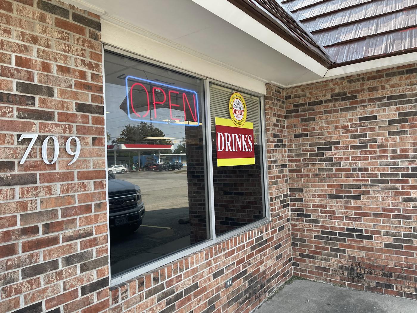 The storefront window at 709 S Maguire St: brick, a neon open sign, the round Yummy's Donut Palace decal and a red and yellow drinks sign.
